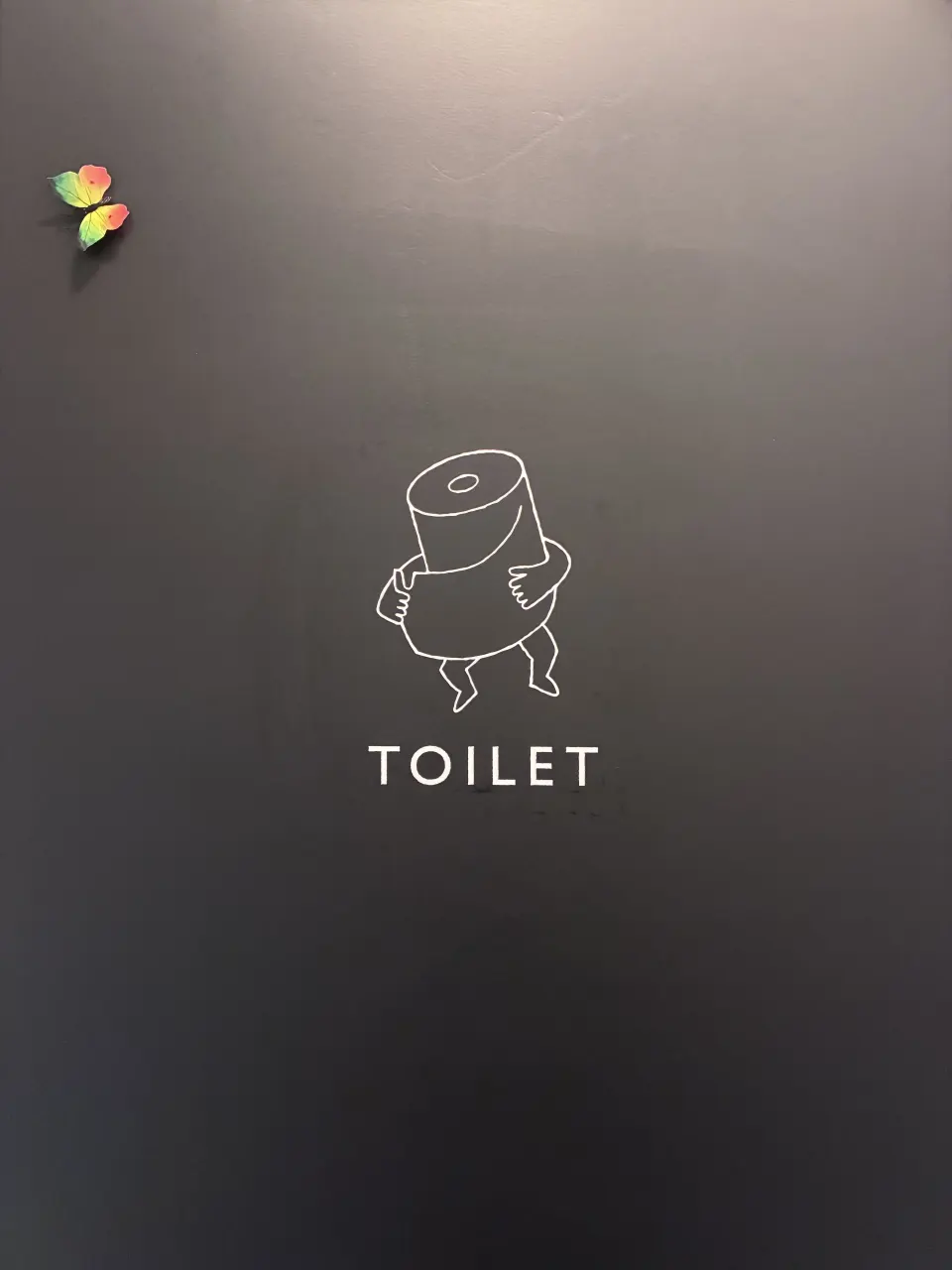 手足の生えたトイレットペーパーが、ほどけた自分の紙を抱えて巻き直しているトイレサイン。黒い扉に白のカッティングシート